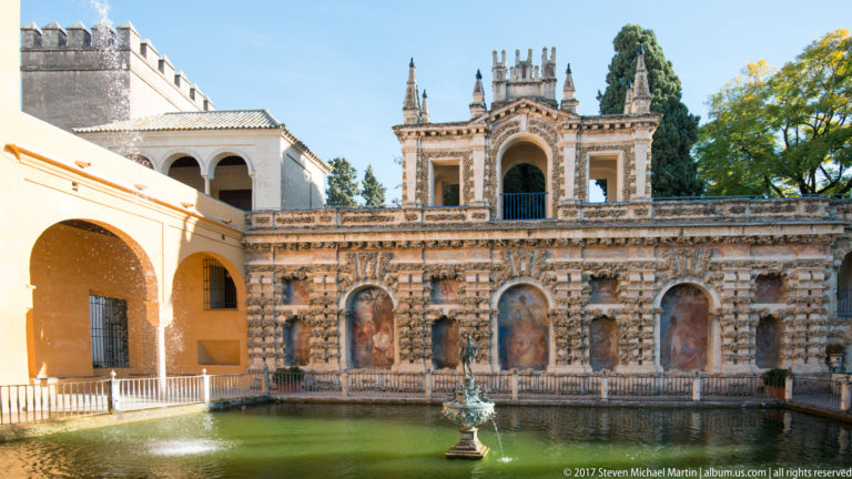 Ornate two-level garden façade with rusticated stonework, arched niches, upper pavilion, and a small fountain in a rectangular pool.