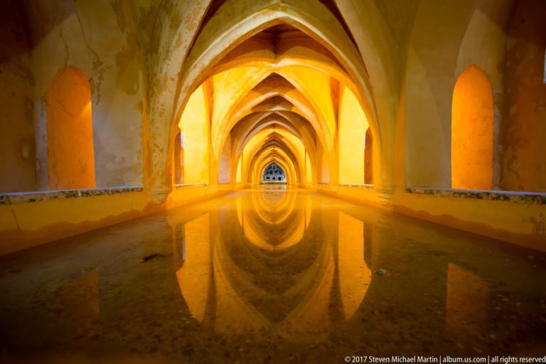 Long vaulted chamber of the Baths of María de Padilla with pointed arches reflected symmetrically in still water and warm side lighting.