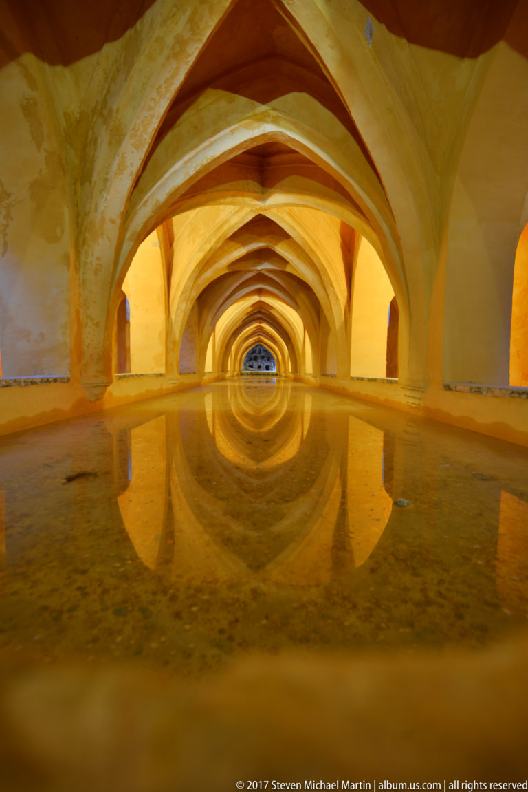 Low waterline view through repeated pointed vaults in the Baths of María de Padilla, with golden arches reflected in still water.