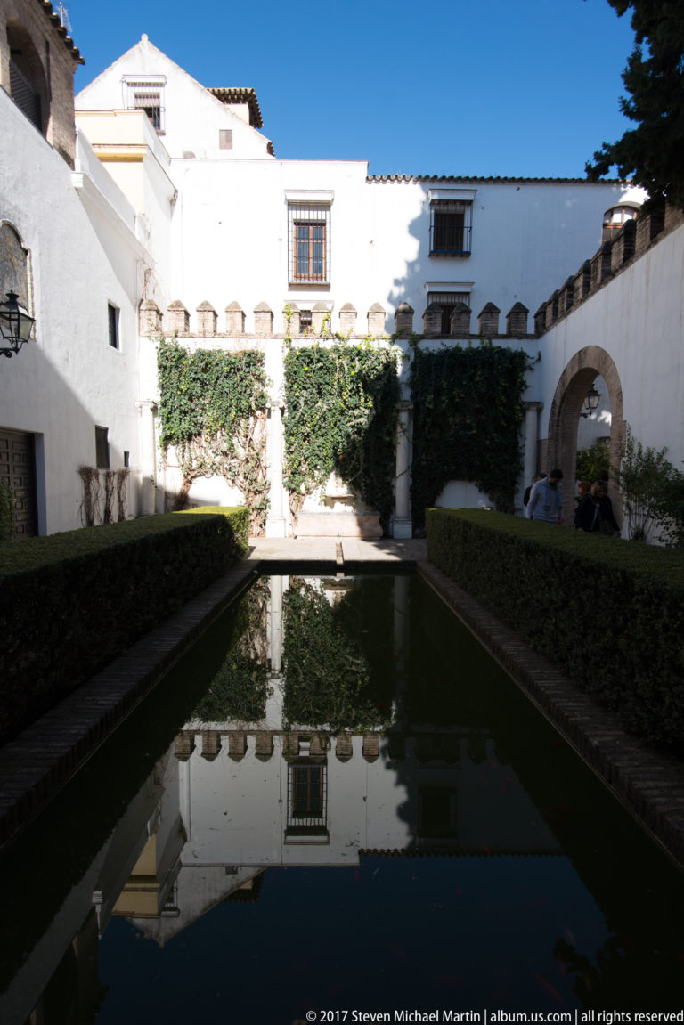 Rectangular reflecting pool bordered by clipped hedges in a white-walled courtyard with ivy, windows, and an arched passage.