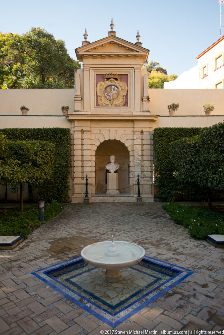Formal garden court with circular fountain basin, blue tile edging, clipped hedges, orange trees, and a pedimented wall fountain.