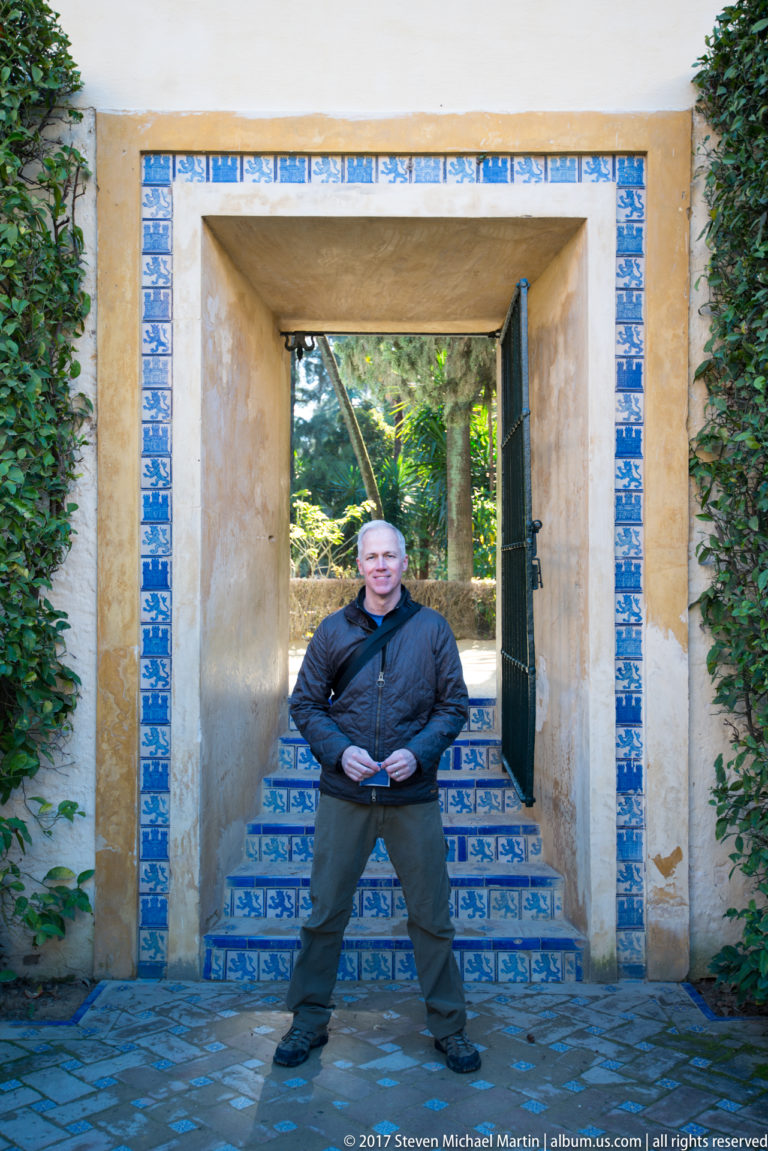 Man standing in a rectangular garden doorway bordered by blue-and-white tiles, with tiled steps and greenery beyond.