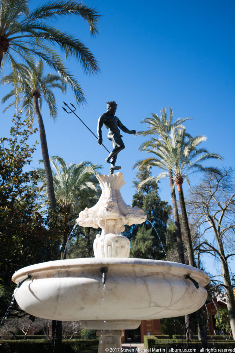 Low-angle view of a garden fountain with dark bronze figure, shell-like stone ornament, broad basin, palms, and blue sky.