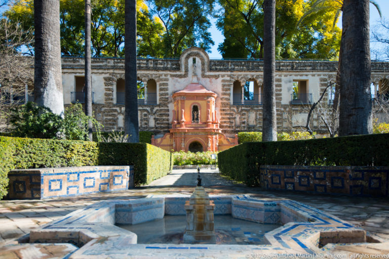 Blue-and-white tiled fountain basin aligned with a path, clipped hedges, palms, and a small red pavilion against a historic wall.