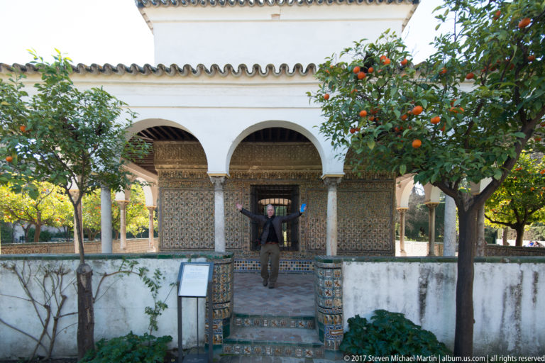White arcaded garden pavilion with patterned tilework, tiled steps, a standing visitor, and an orange tree bearing fruit.