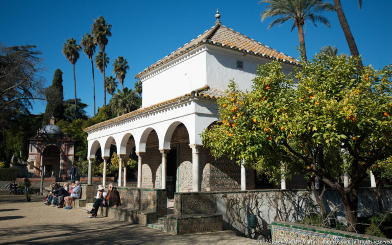 White garden pavilion with rounded arcade, patterned tile panels, orange trees, palms, and seated visitors.