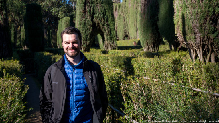 Man facing the camera on a narrow garden path bordered by clipped hedges and tall cypress trees.