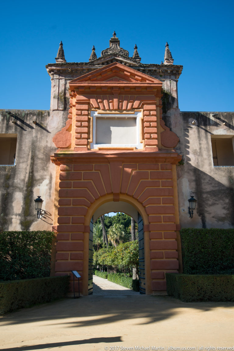 Red-and-ochre rusticated gateway with a large central arch, decorative upper section, clipped hedges, and garden visible beyond.
