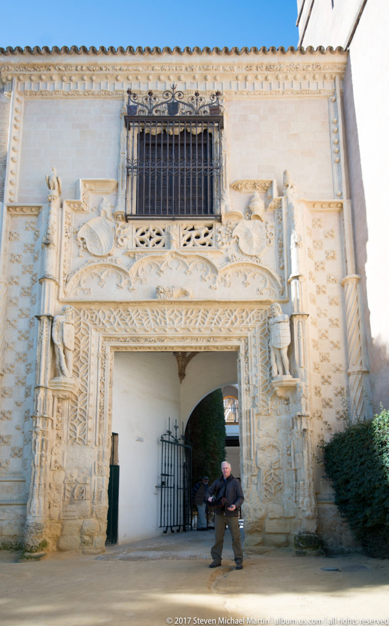 Ornately carved pale-stone gateway with relief decoration, heraldic shields, barred upper window, and a visitor walking beneath the arch.