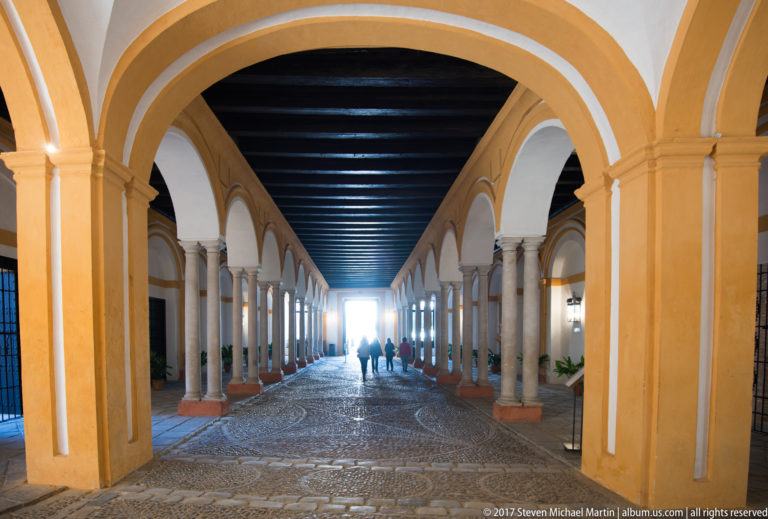 Long gallery with yellow-and-white arches on stone columns, dark timber ceiling, patterned paving, and visitors in the distance.