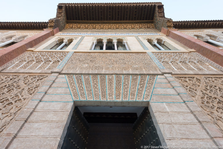 Upward view of the Palace of Pedro I entrance with stone blocks, blue-green tile bands, carved stucco, windows, and timber eaves.