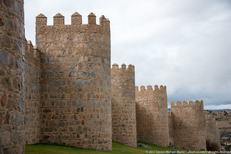 Close view of crenellated stone towers projecting from Ávila’s medieval city wall above a grassy base.