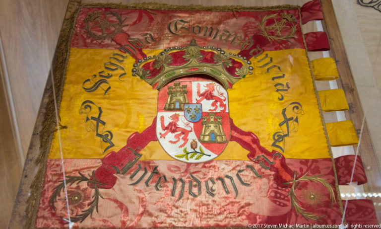 Aged yellow and red embroidered banner with a crowned coat of arms, cursive lettering and decorative fringe.