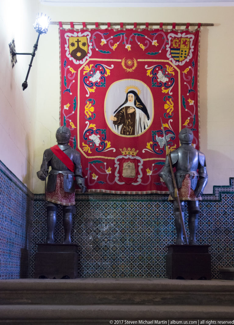 Two armored figures flank a red heraldic tapestry with a central portrait medallion in a museum room.
