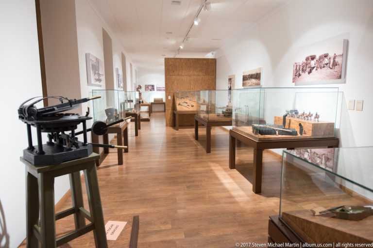 Museum gallery with glass display cases, historical objects, wall panels and a large metal apparatus at left.