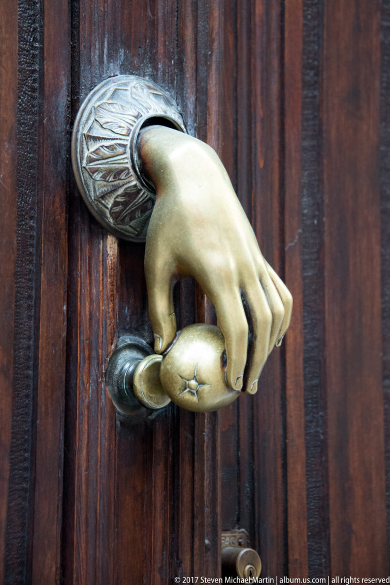 Brass hand-shaped door knocker gripping a rounded striker on a dark vertically grained wooden door.
