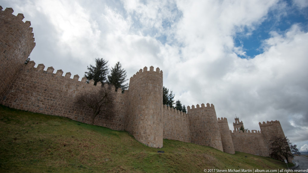 Stone crenellated walls and projecting towers rise along a grassy slope beneath a cloudy sky.
