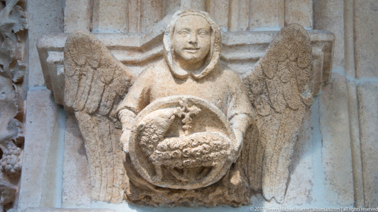 Weathered carved stone winged figure holding a circular wreath or medallion against Gothic architectural stonework.