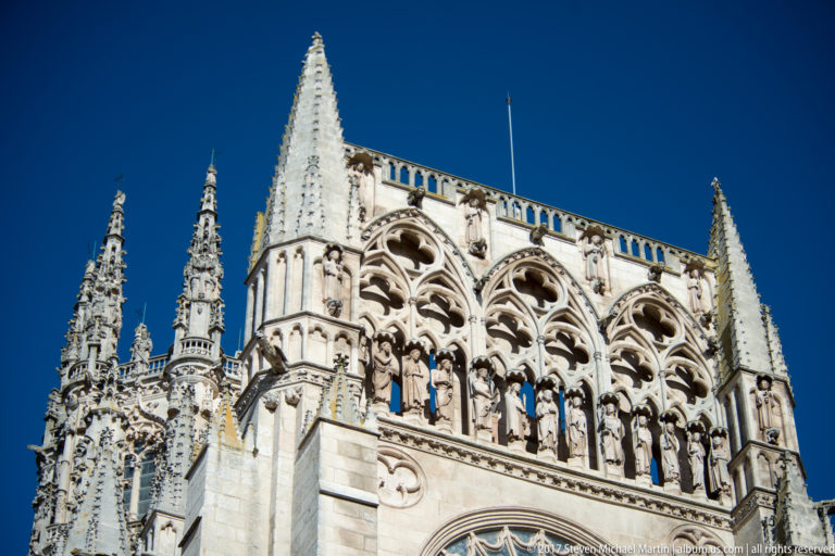 Upward view of Burgos Cathedral with pinnacles, open Gothic tracery, sculpted figures, and pale stonework against blue sky.
