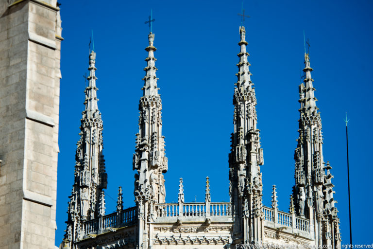 Four ornate Gothic stone pinnacles and roof balustrade of Burgos Cathedral against a cloudless blue sky.
