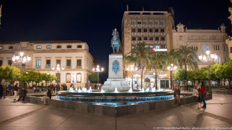 Equestrian Gran Capitán monument stands above a lit fountain in Plaza de las Tendillas, surrounded by pedestrians and city buildings.