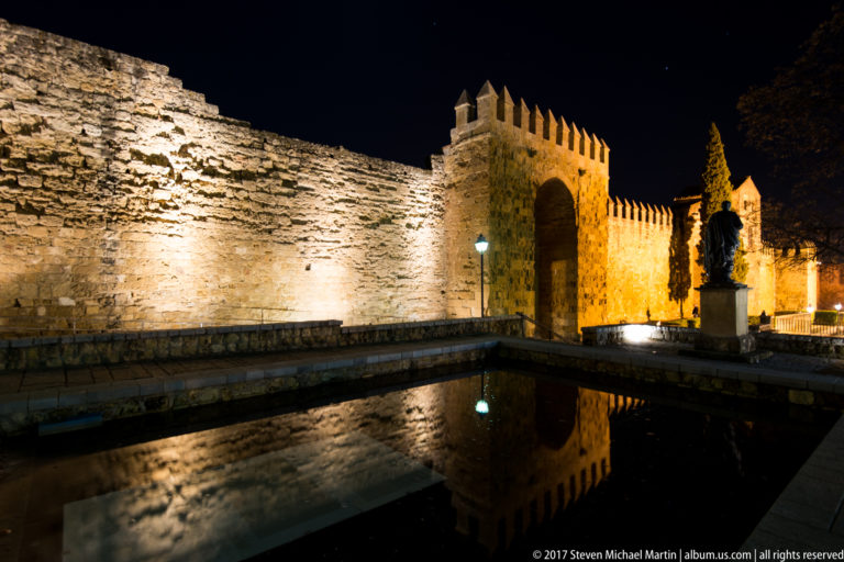 Illuminated crenellated gateway and wall reflected in a rectangular pool, with a standing monument at right.