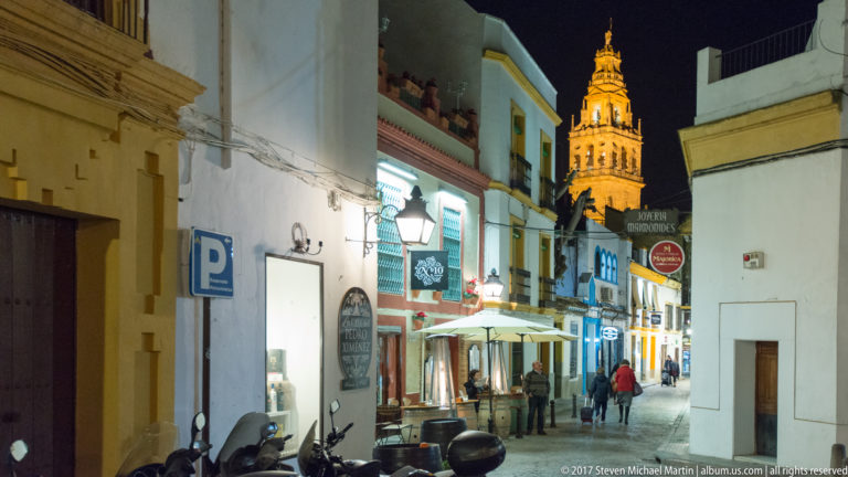 Narrow Córdoba street with cafés, scooters, pedestrians, and the illuminated Mosque-Cathedral bell tower rising in the distance.