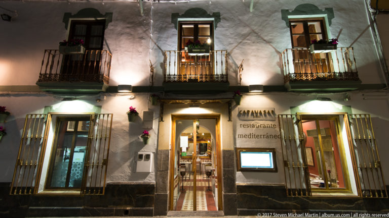 Whitewashed restaurant façade with barred windows, small balconies, flower boxes, and an open central doorway at night.