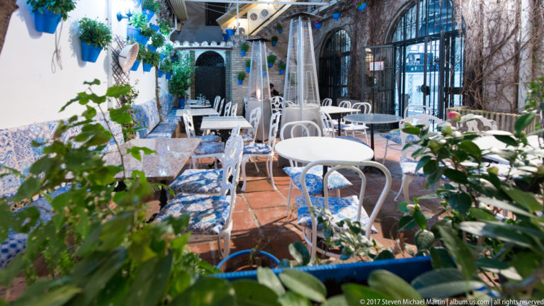 Plant-filled restaurant courtyard with white metal tables and chairs, blue patterned tile benches, and terracotta flooring.