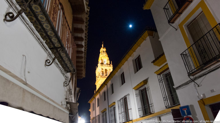 Illuminated Mosque-Cathedral bell tower framed by white buildings and balconies on a narrow street beneath the moon.