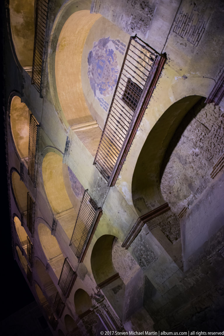 Steeply angled view of an old stone-and-plaster arcade with iron balconies, repeated arches, and faded circular painted medallions.