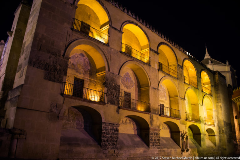 Warmly illuminated two-story arcade with repeated semicircular arches, iron balconies, exposed stone, and faded painted medallions.