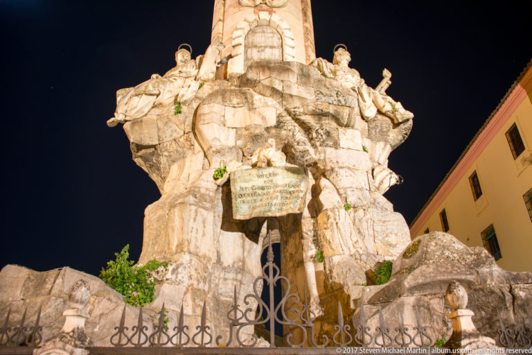 Floodlit Baroque monument base formed from sculpted rockwork and figures, with a central inscription and iron fence.