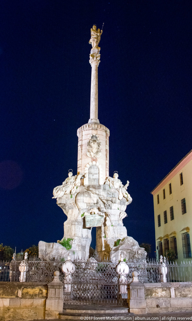 Illuminated Saint Raphael triumph monument with a tall column, sculptural base, iron enclosure, and historic buildings behind.