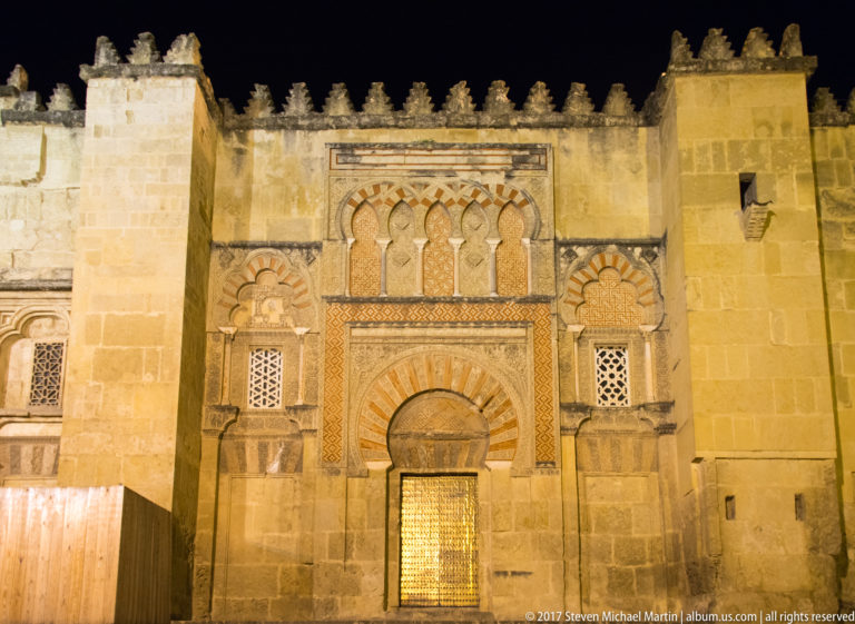 Illuminated Mosque-Cathedral doorway with striped arch, interlaced blind arches, carved panels, lattice windows, and crenellated roofline.