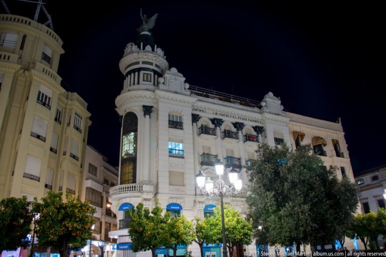 White corner building with rounded turret, balconies, elaborate roofline, and rooftop figure above orange trees at night.