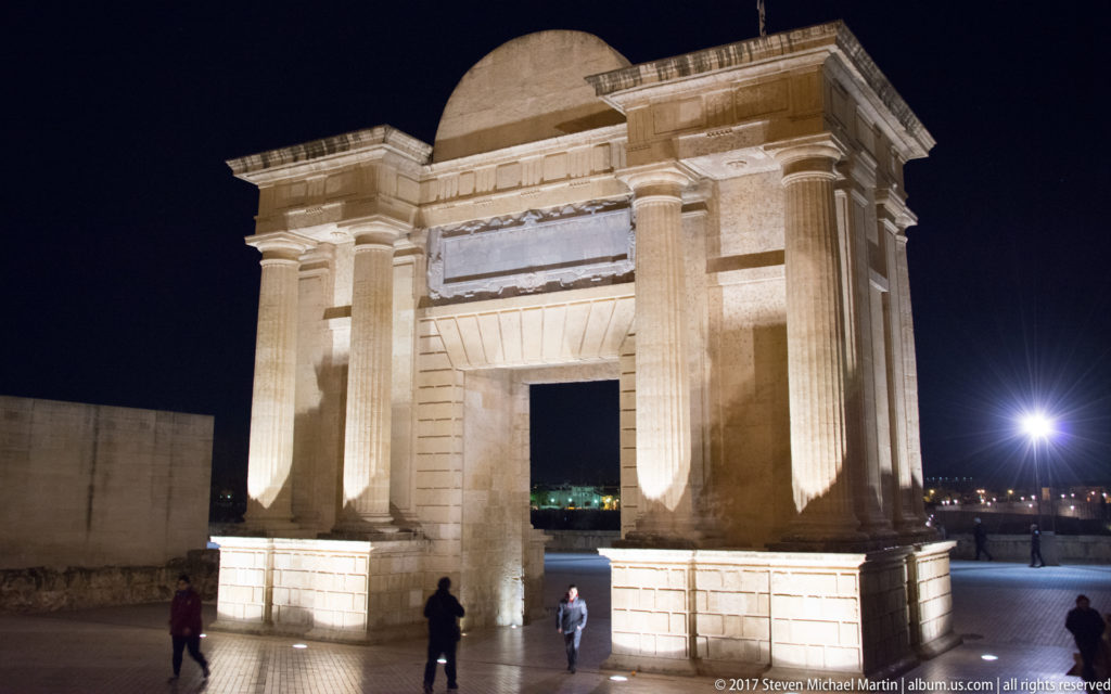 Floodlit Puerta del Puente with paired classical columns and central opening, seen at night with pedestrians at its base.