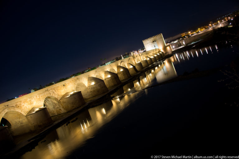 Diagonal nighttime view of the illuminated Roman Bridge reflected in the Guadalquivir, with Calahorra Tower at the far end.