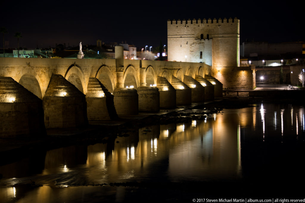 Illuminated Roman Bridge arches reflected in the Guadalquivir, with the square Calahorra Tower rising at the far end.