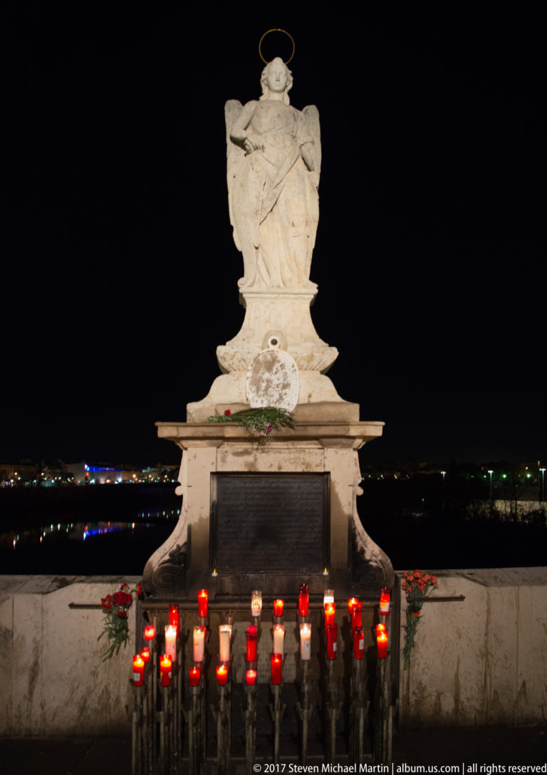 White statue of Saint Raphael with halo above a pedestal lined with red votive candles and flowers on the Roman Bridge.