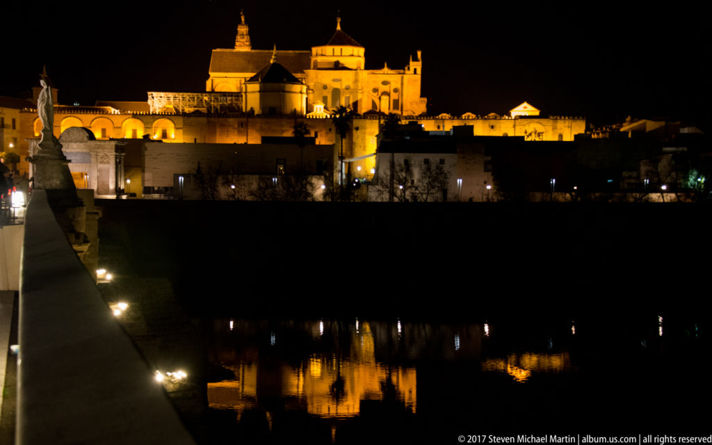Illuminated Mosque-Cathedral skyline across the Guadalquivir, with reflections and part of the Roman Bridge in the foreground.