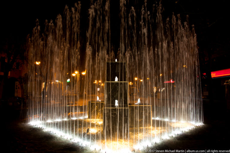 Brightly illuminated circular fountain with dense vertical water jets against a dark Córdoba streetscape at night.