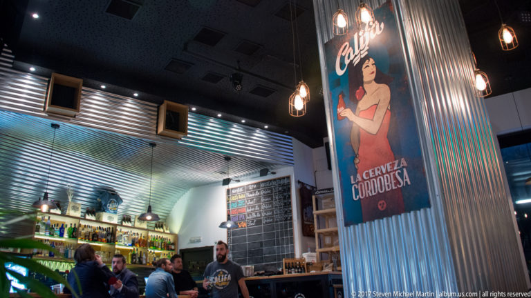 Califa beer bar interior with patrons, counter, chalkboard menu, hanging bulbs, and a poster reading La cerveza cordobesa.