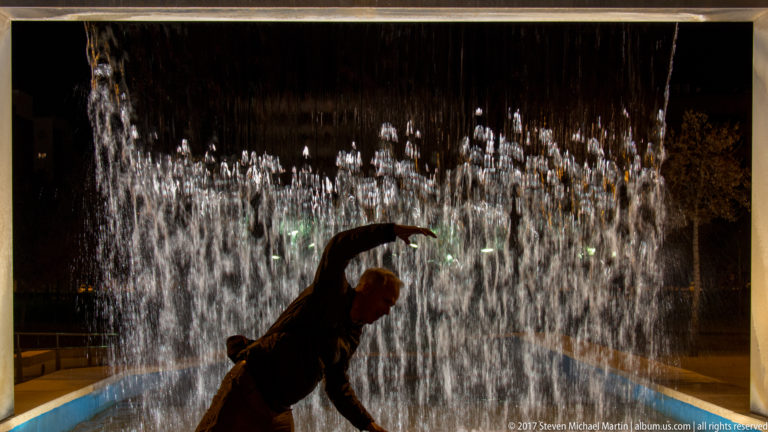 Silhouetted person bends through a curtain of bright vertical fountain jets at night in Córdoba.