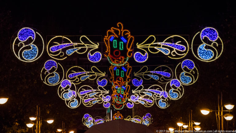 Multiple rows of colorful carnival lights form a receding canopy of ornate scrollwork and mask motifs above a Córdoba street.