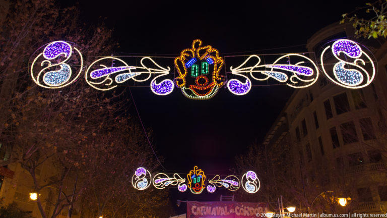 Colorful carnival light arches with mask motifs hang above a Córdoba street, with a Carnaval de Córdoba banner below.
