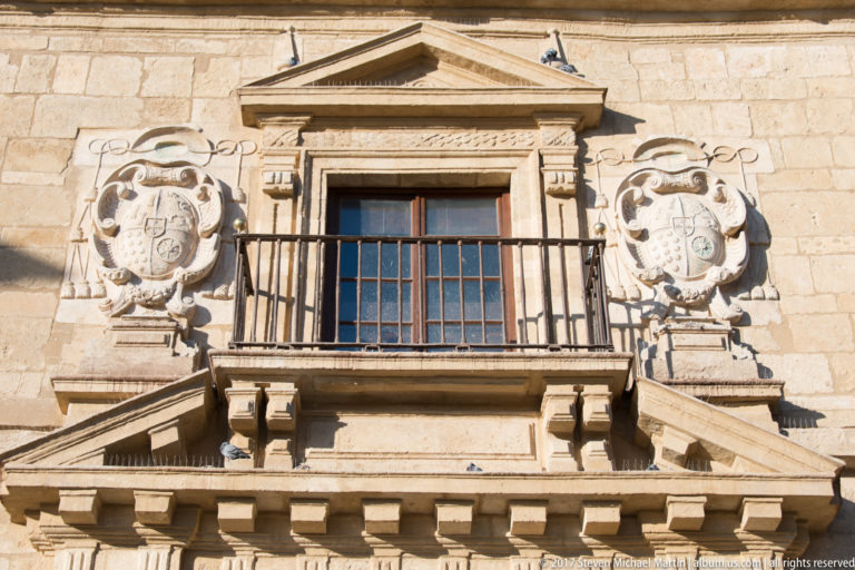Carved stone façade with central iron balcony, triangular pediment, pilasters, corbels, and large heraldic shields on both sides.