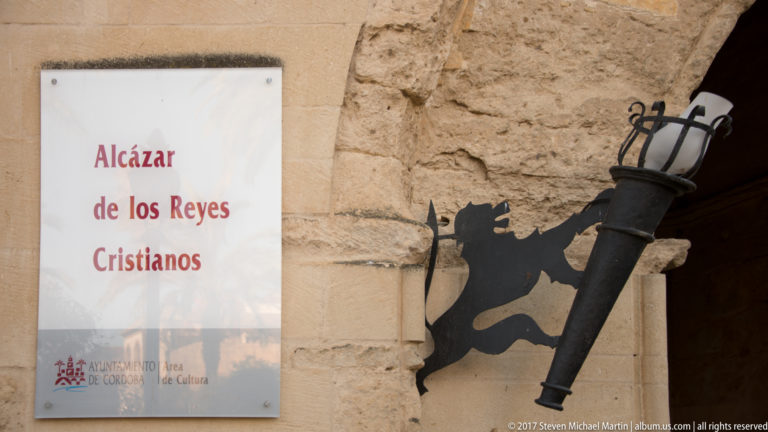 Entrance plaque reading Alcázar de los Reyes Cristianos beside a black metal lantern bracket shaped like a lion.