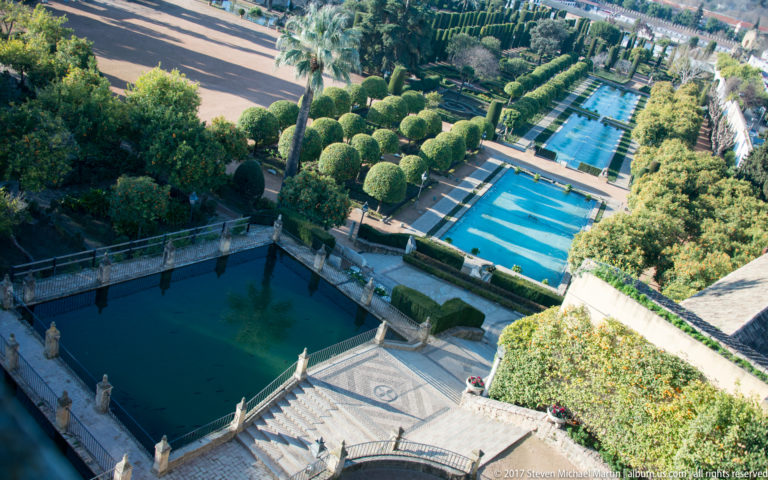 Elevated view over rectangular pools, clipped hedges, rounded topiary, palms, and terraces in the Alcázar gardens.