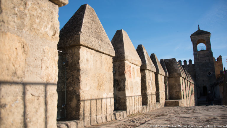 Low-angle view along the pointed crenellations of the Alcázar wall with a small tower rising behind.